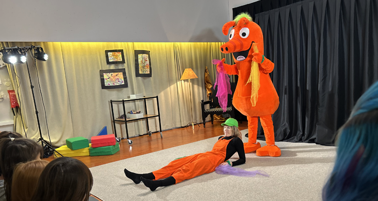 Bild från föreställningen "Språkfiluren leker" där den orangea maskoten Språkfiluren och en kompis leker på en scen.