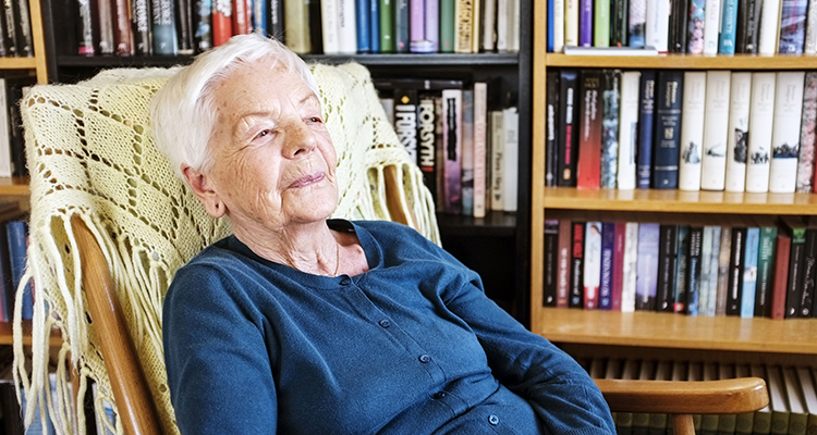 En äldre kvinna sitter i fåtölj framför en bokhylla.