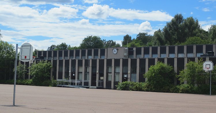 Asfalterad skolgård med basketkorg placerad i varsitt hörn, i bakgrunden en vit och svart randig byggnad med en klocka placerad i mitten av fasaden 