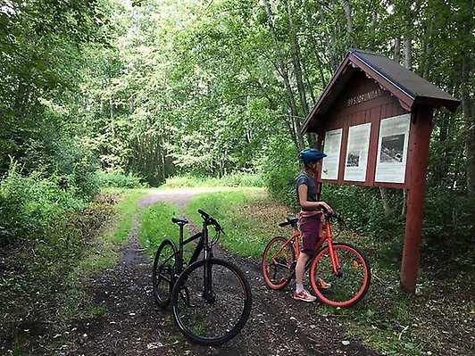 Cyklar på skogsväg intill informationsskylt Bysjön. Foto.